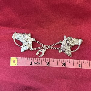 May include: Silver-toned horse head brooch with a horseshoe charm. The brooch features two detailed horse heads connected by a chain, with a horseshoe in the center. The brooch measures approximately 4 inches in length.