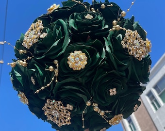May include: A dark green bouquet of silk roses with gold accents and sparkling rhinestones. The bouquet is held in the air against a blue sky.