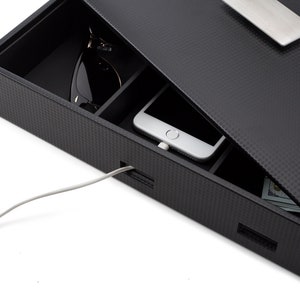 May include: A black charging station with a carbon fiber pattern and a silver handle. The station has compartments for a phone, sunglasses, and cash. A white charging cable is plugged into the station.