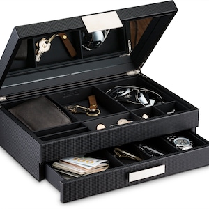 May include: A black, multi-compartment valet box with a carbon fiber pattern. The open box reveals a mirrored lid, sunglasses, a wallet, keys, coins, a watch, and a drawer with cash. The box has a silver-colored handle.