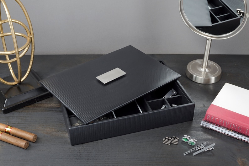 May include: A black leather jewelry box with a silver metal accent on the lid. The box is open and has compartments for storing jewelry. The box is on a dark wood surface.