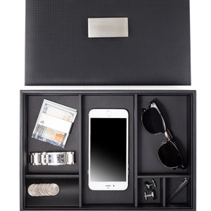 May include: A black valet tray with compartments for a phone, sunglasses, watch, money, cufflinks, and a pen. The tray has a textured surface and a silver metal nameplate.