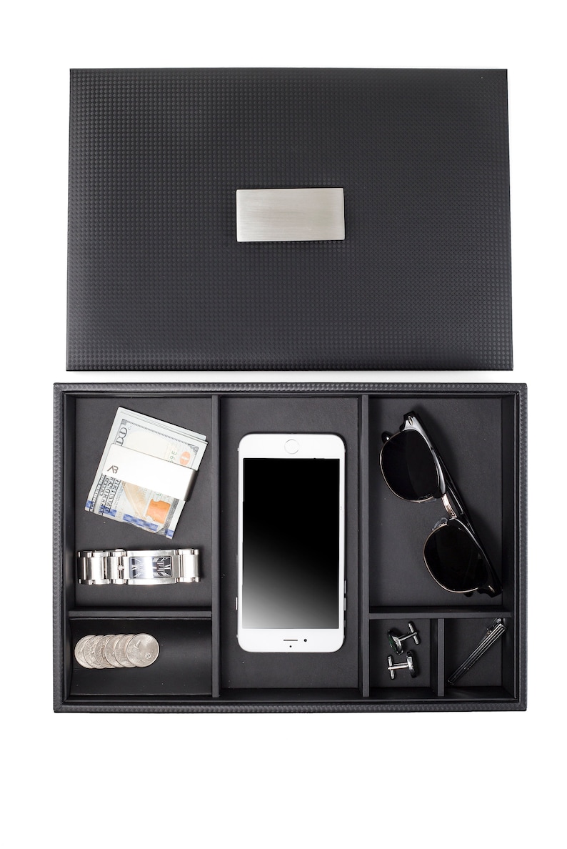 May include: A black valet tray with compartments for a phone, sunglasses, watch, money, cufflinks, and a pen. The tray has a textured surface and a silver metal nameplate.