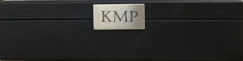 May include: A black rectangular box with a silver metal plate on the top. The plate has the letters "KMP" engraved on it.