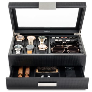 May include: Black watch and accessory organizer with a glass top. The organizer holds six watches, sunglasses, cufflinks, and other accessories. The bottom drawer contains cigars, a brush, and other items. The organizer is made of a textured material.