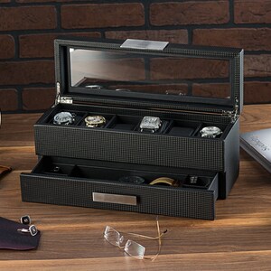 May include: A black watch box with a glass top and a drawer. The box has a grid pattern and is open to reveal several watch compartments. There are watches in the compartments.