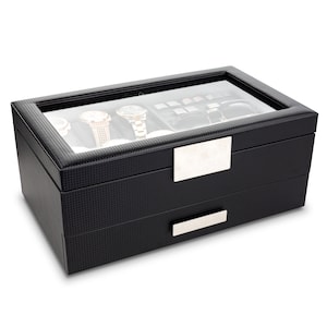 May include: A black watch display case with a carbon fiber texture. The top section has a glass lid, showcasing several watches. The bottom section has a drawer with a silver handle. The case has a rectangular silver plate on the front.