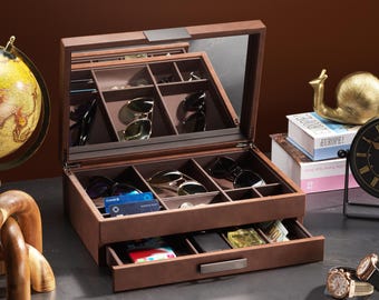 May include: A brown leatherette sunglasses and accessories organizer with a mirrored lid. The open organizer reveals multiple compartments holding sunglasses, credit cards, and other items. A drawer is partially open, displaying additional storage.