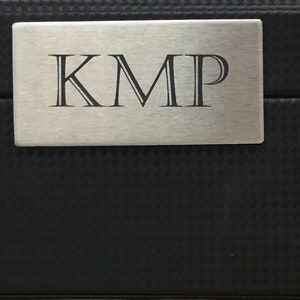 May include: A black rectangular box with a silver metal plate on the top. The plate has the letters "KMP" engraved on it.