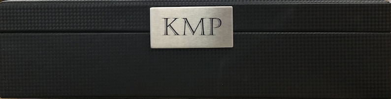 May include: A black rectangular box with a silver metal plate on the top. The plate has the letters "KMP" engraved on it.