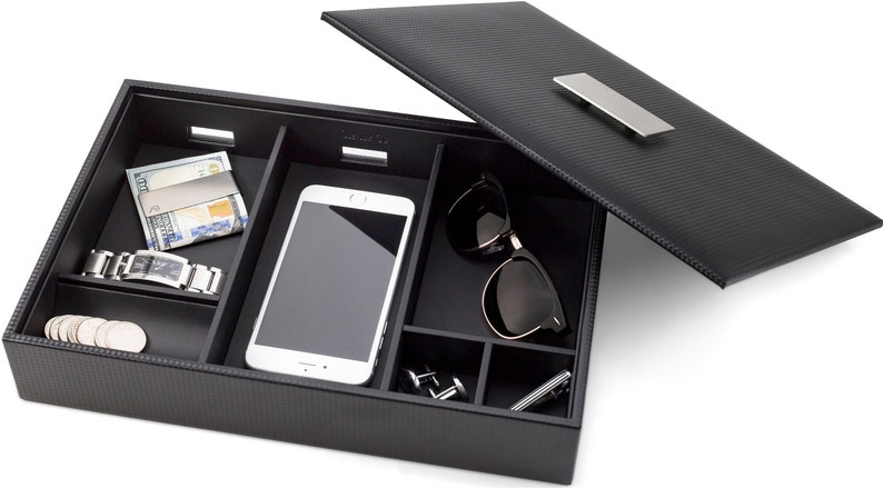 May include: A black leather valet tray with compartments for organizing items like a phone, sunglasses, watch, and cufflinks. The tray has a textured, carbon fiber-like finish and a silver metal handle.