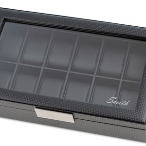 May include: A black watch box with a glass top and 12 compartments. The box has a carbon fibre pattern and the word "Smith" is engraved on the front.