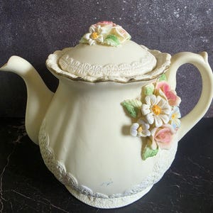May include: A cream-coloured ceramic teapot featuring a lid topped with sculpted flowers. The body is decorated with embossed lace-like patterns at the base and rim, and a cluster of three-dimensional pink, white, and blue flowers on the side.
