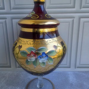 May include: A deep red and gold glass jar with a matching lid, featuring a hand-painted floral design with red and blue flowers and green leaves. The jar has a wide, rounded body, a narrow neck, and a clear glass stem base with a gold-toned ring.