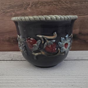 May include: A dark charcoal ceramic planter featuring a raised relief design of red berries and blue flowers around the exterior. The rim has a textured rope-like pattern, and the overall finish is glossy, highlighting the detailed botanical motifs.