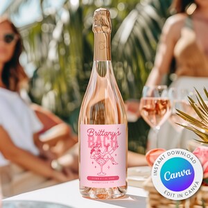 Puede incluir: Una botella de champán rosado con una tapa de lámina dorada. La etiqueta dice "Brittany's Bach" con una ilustración rosa de una mujer en una copa de martini. La botella está sobre una mesa blanca con dos copas de champán rosado.