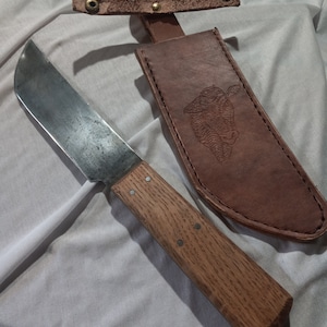 May include: A brown leather sheath with a cow head design and a metal snap closure. A long, thin, metal knife with a wooden handle is lying on a white fabric surface.
