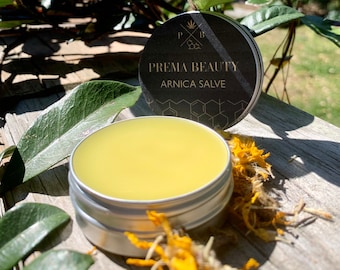 May include: A small silver tin of Arnica Salve with a black label that reads "Prema Beauty Arnica Salve". The tin is sitting on a wooden surface with yellow flowers.