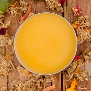 May include: A round container of yellow balm on a wooden surface with dried flowers and leaves scattered around it.