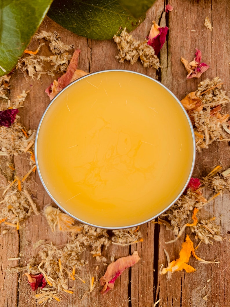 May include: A round container of yellow balm on a wooden surface with dried flowers and leaves scattered around it.