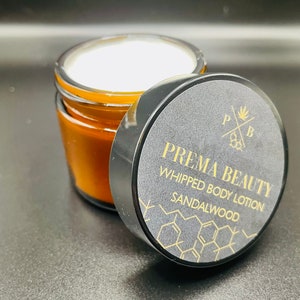 May include: A black jar with a gold lid containing a white whipped body lotion. The lid has the text "PREMA BEAUTY" and "WHIPPED BODY LOTION" in gold lettering. The text "SANDALWOOD" is also in gold lettering.