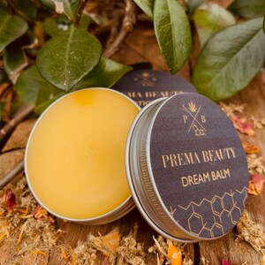 May include: A small tin of yellow balm with a black lid. The lid has a gold logo that says "Prema Beauty Dream Balm" with a honeycomb pattern around the text.
