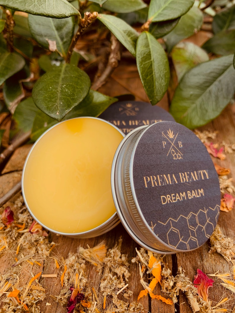 May include: A small tin of yellow balm with a black lid. The lid has a gold logo that says "Prema Beauty Dream Balm" with a honeycomb pattern around the text.