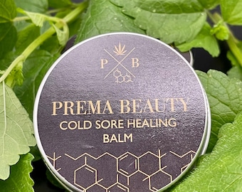 May include: A round, silver-rimmed tin of Prema Beauty Cold Sore Healing Balm, set against a backdrop of green leaves. The tin has a black label with gold text and a geometric pattern. The Prema Beauty logo is at the top.