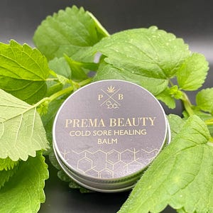Puede incluir: Una pequeña lata plateada con una etiqueta negra que dice "Prema Beauty Cold Sore Healing Balm". La etiqueta tiene un diseño de hoja de cannabis dorada y un patrón de panal. La lata está sobre un lecho de hojas verdes.