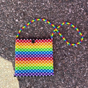 May include: A square-shaped handbag constructed from small, multi-colored plastic beads arranged in a vibrant rainbow-striped pattern. The bag features a long, matching rainbow-beaded shoulder strap and a single black bead at the top center.
