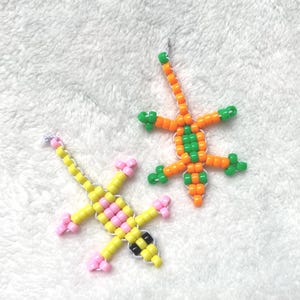 以下が含まれることがあります： 2つのビーズで作られたトカゲの置物。1つはピンクと黒のアクセントが付いた黄色、もう1つはオレンジと緑です。トカゲは、小さなカラフルなビーズを繋ぎ合わせて作られており、白いテクスチャのある表面に置かれています。