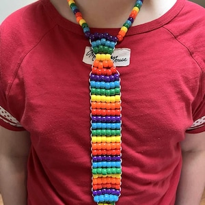 Könnte beinhalten: Eine farbenfrohe Krawattenkette aus Perlen in Regenbogenfarben, darunter Rot, Orange, Gelb, Grün, Blau und Lila. Die Krawatte besteht aus kleinen, runden Perlen, die aneinandergereiht sind. Die Halskette wird um den Hals getragen.