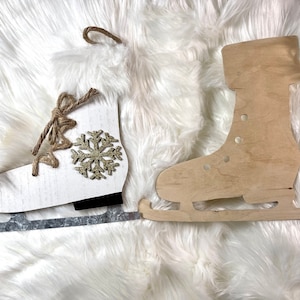 May include: Two wooden ice skate ornaments, one painted white with a gold snowflake and the other in natural wood, on a white fluffy surface.