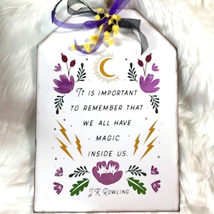 Puede incluir: Un letrero de madera blanco con una cita de J.K. Rowling: "Es importante recordar que todos tenemos magia dentro de nosotros." El letrero está decorado con una luna creciente, estrellas, flores y rayos en morado, dorado y verde.