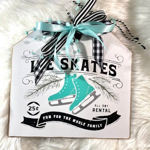 May include: White wooden sign with black and white lettering that says "Ice Skates" with a graphic of two teal ice skates. The sign also says "Fun for the whole family" and "All Day Rental" with a price of 25 cents. The sign is decorated with a black and white checkered ribbon and a light blue ribbon with white pom-poms.