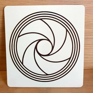 May include: A square, off-white wooden plaque featuring a dark brown, stylized aperture design. The design consists of a central circle surrounded by concentric rings and curved, blade-like shapes radiating outwards. The plaque is set against a light wood surface.