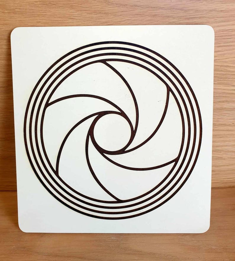 May include: A square, off-white wooden plaque featuring a dark brown, stylized aperture design. The design consists of a central circle surrounded by concentric rings and curved, blade-like shapes radiating outwards. The plaque is set against a light wood surface.