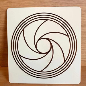 May include: A square, off-white wooden plaque featuring a brown, stylized aperture design. The design consists of a central circle surrounded by curved blades and concentric rings, creating a visual representation of a camera lens.