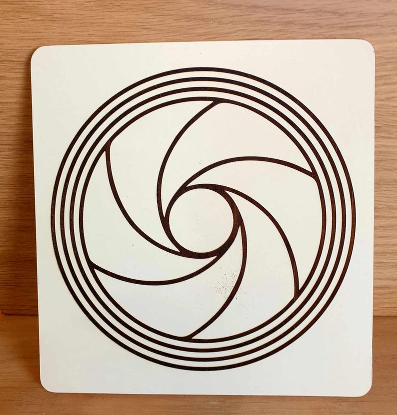 May include: A square, off-white wooden plaque featuring a brown, stylized aperture design. The design consists of a central circle surrounded by curved blades and concentric rings, creating a visual representation of a camera lens.