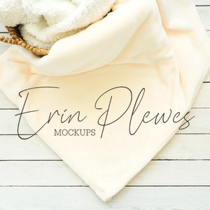Pode incluir: Um cobertor macio de cor creme drapeado sobre uma cesta de vime em um fundo de madeira branca. O texto "Erin Plewes Mockups" está impresso no cobertor.
