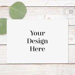 Puede incluir: Una tarjeta blanca con el texto "Your Design Here" sobre un fondo de madera blanqueada con hojas de eucalipto.