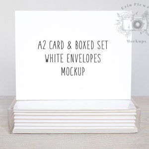 May include: A white card and envelope set mockup. The card is blank and the envelopes are stacked in a clear plastic box. The text on the card reads "A2 Card & Boxed Set White Envelopes Mockup".