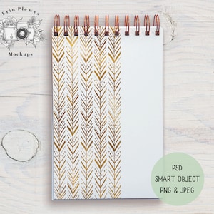 Pode incluir: Um caderno branco em espiral com um padrão de chevron dourado no lado esquerdo. O caderno está sobre uma superfície de madeira branca. PSD SMART OBJECT PNG & JPEG