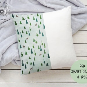 Puede incluir: Almohada blanca con un estampado de bosque verde y marrón en un lado. La almohada está sobre una superficie de madera blanca con una manta gris.