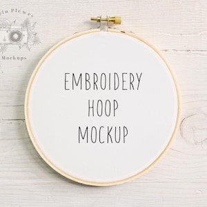 Puede incluir: Maqueta de aro de bordado blanco sobre un fondo de madera blanca. El aro tiene el texto "Embroidery Hoop Mockup" escrito en él.