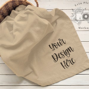May include: A beige blanket with the text "Your Design Here" is draped over a woven basket. The blanket is folded and has a soft texture. The basket is made of woven straw and sits on a white wooden surface.