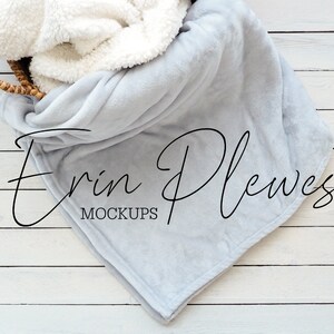 May include: A soft, light gray throw blanket with a white sherpa lining is draped over a woven basket on a white wood background. The text "Erin Plewes Mockups" is printed on the blanket.