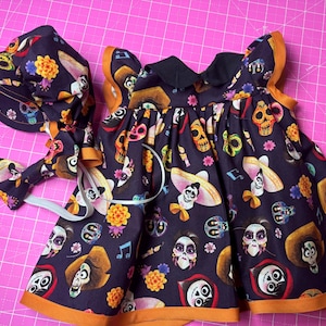 May include: A dark navy blue baby dress and matching bonnet set. The fabric features a colorful print of sugar skulls, sombreros, and musical notes. The dress has orange trim and a black collar.