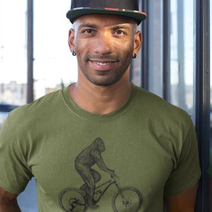 Puede incluir: Un hombre con una gorra de b&eacute;isbol negra con una franja roja, blanca y azul lleva una camiseta verde con una ilustraci&oacute;n en blanco y negro de un gorila montando en bicicleta.
