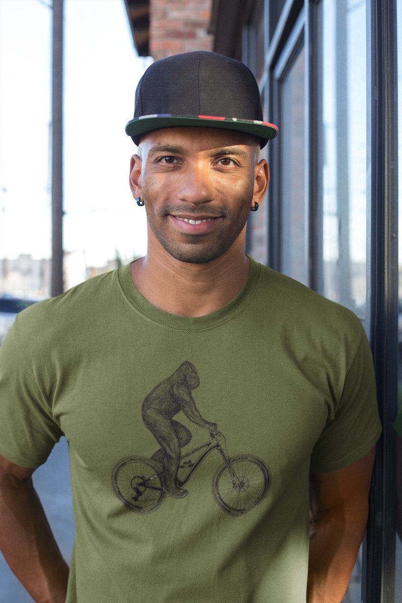 Puede incluir: Un hombre con una gorra de b&eacute;isbol negra con una franja roja, blanca y azul lleva una camiseta verde con una ilustraci&oacute;n en blanco y negro de un gorila montando en bicicleta.